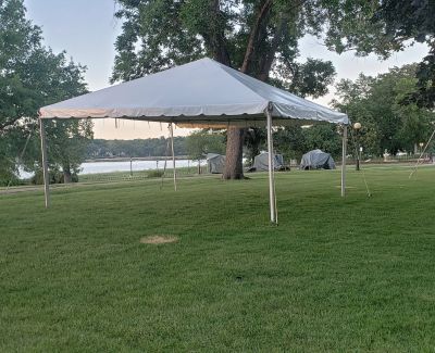 A single white frame tent set up on a grassy slope overlooking a body of water, with trees framing the scene and other tents visible in the distance.