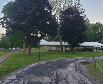 A cracked asphalt path winding through a park with large trees, leading towards a long white event tent in the background.