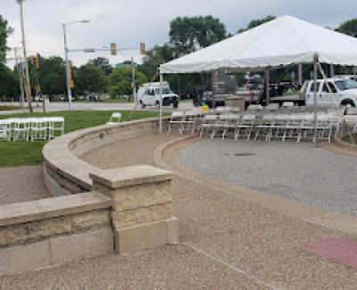 View from behind a curved stone retaining wall looking towards a large white tent set up with rows of white folding chairs on a paved area, near a street intersection.