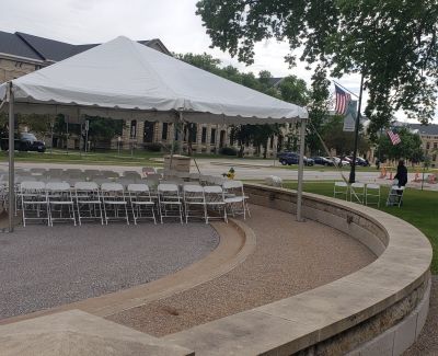 Rows of white folding chairs set up under a white event tent situated along a curved, low stone wall, with a building and trees in the background.