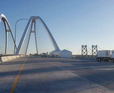 View along the roadway of a modern cable-stayed bridge with large white arches, showing white tents and a utility truck set up on the bridge deck under a clear blue sky, with an older truss bridge section visible further down.