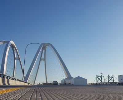 Low angle view of the large, white, arched structure of a modern cable-stayed bridge against a clear blue sky. Tents and an older truss bridge section are visible further down the bridge deck.
