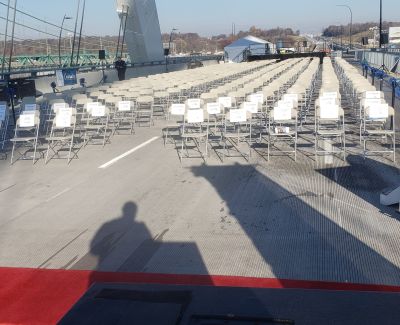 Rows of empty white folding chairs arranged on a bridge deck facing away from the camera, with a red carpet runner down the center aisle. The photographer's shadow is cast on the pavement.