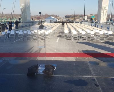 View down the red carpet aisle between rows of empty white chairs set up for an event on a bridge deck, leading towards a podium and the bridge structure in the distance.