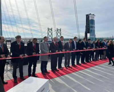Group of officials in suits lined up on a red carpet, cutting a red ribbon for a ceremony, likely opening the large bridge visible in the background.