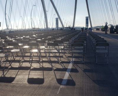 Rows of empty grey folding chairs casting long shadows on the bridge deck under bright sunlight, beneath the cables of the bridge structure.
