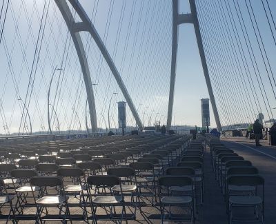 Numerous rows of empty grey folding chairs arranged on the deck of a cable-stayed bridge, seen from behind, with the bridge's large white arches and cables prominent against the sky.