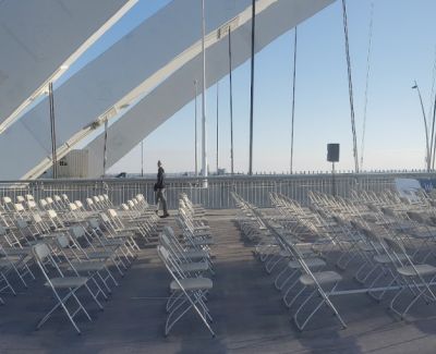 View from beneath the angled white support structure and suspension cables of the bridge, looking over rows of empty white folding chairs set up on the deck.