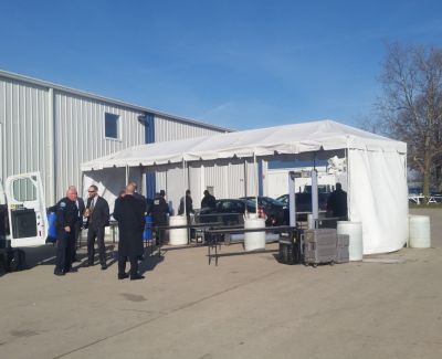 People standing outside under and near a white frame tent set up on pavement next to a large, light-colored industrial building under a clear blue sky.