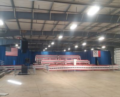 Interior view of a large, brightly lit hall or warehouse set up for an event, featuring tiered bleachers decorated with patriotic bunting and an American flag on the stage backdrop.