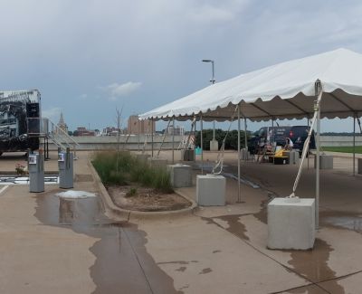 A large white event tent secured with heavy concrete blocks on a paved outdoor area, with wet patches on the ground.