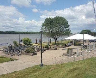 Panoramic view of a riverside park area with tiered concrete seating, overlooking a wide river under a partly cloudy sky. A white tent is set up on the lower level.