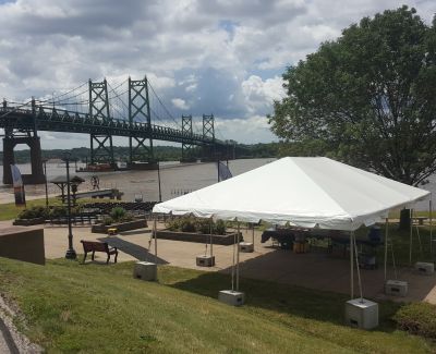 A white event tent set up on a grassy knoll overlooking a river, with a large green truss bridge spanning the water in the background.