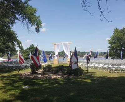 Outdoor wedding ceremony setup on a lawn by a river, with rows of white chairs facing a draped archway, flanked by various flags under a blue sky.