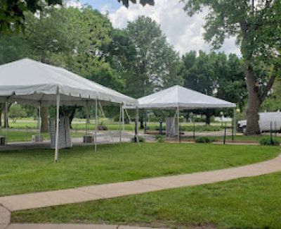 Two white canopy tents set up on a grassy area in a park, next to a winding paved path and surrounded by trees.