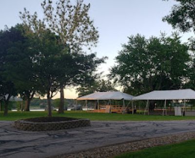 Park setting at dusk featuring a paved area with a stone planter, leading to a grassy area with several white event tents near trees and overlooking water.