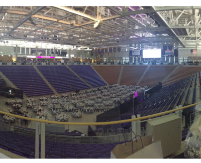 An elevated view inside a large sports arena, showing the arena floor set with numerous round tables dressed in white for a banquet or event, surrounded by tiers of purple and orange stadium seating.