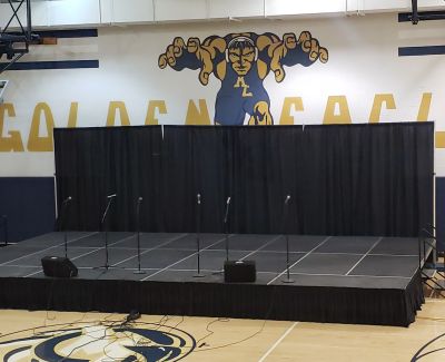 A portable stage with black skirting and a black pipe-and-drape backdrop set up on a gymnasium floor, equipped with microphones and speakers, positioned below a large 