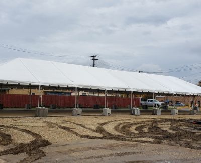 A very long white frame tent installed on a dirt lot, heavily weighted down with numerous concrete blocks along its perimeter, with a fence visible behind it under an overcast sky.