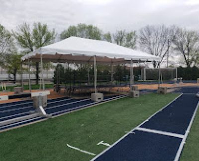 A white frame tent with a partially clear roof covering an outdoor patio area with fixed benches and tables, possibly adjacent to a running track or sports field, secured with concrete weights.