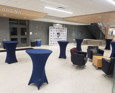 The modern lobby or pre-function area of an arena, featuring geometric tiled flooring, several high-top cocktail tables with blue spandex covers, and a comfortable lounge seating arrangement.