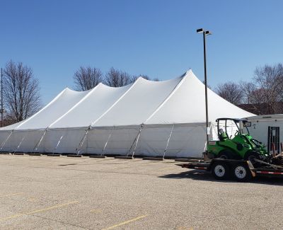 A large multi-peak white Century-style tent pitched in an asphalt parking lot on a clear day, with a green utility vehicle on a trailer parked nearby.