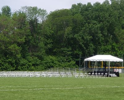 An outdoor event setup on a green field, with rows of white folding chairs facing a small stage covered by a white frame tent against a backdrop of lush green trees.