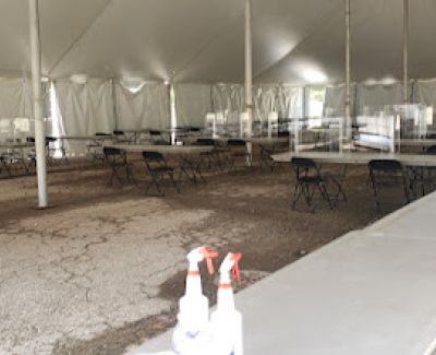 Interior view of a large tent with a gravel floor, sparsely furnished with tables and chairs. Two cleaning spray bottles sit on a table in the foreground.