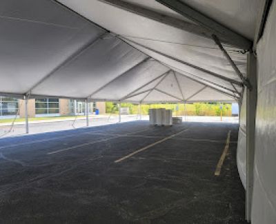 View from inside a large white frame tent looking out onto an empty asphalt parking lot, with stacked white items visible in the distance.