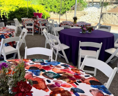 Outdoor event seating with round tables set with colorful patterned tablecloths and solid purple tablecloths, white folding chairs, and floral centerpieces.