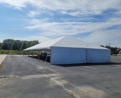 A long white frame tent with solid white sidewalls installed in an asphalt parking lot under a partly cloudy sky.
