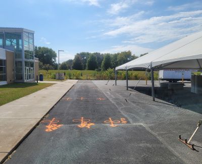 An asphalt area marked with multiple orange spray-painted symbols, adjacent to a modern building, with a large white frame tent covering part of the marked space.