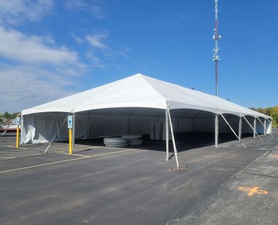 A large white frame tent being installed or standing in an asphalt parking lot, secured with stakes, under a bright blue sky with a communication tower visible.