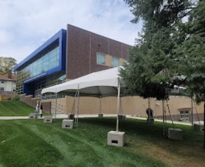 A small white frame tent erected on a grassy area in front of a modern multi-story building, secured with concrete block weights.