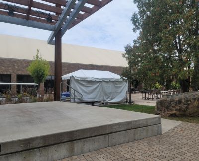 A small tent frame covered with white tarps sitting on a permanent concrete outdoor stage beneath a modern pergola structure.