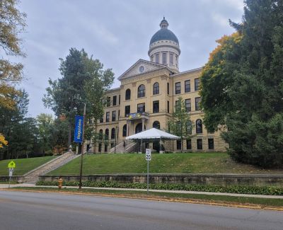 View from across the street of a large, multi-story stone building with a dome on the Augustana College campus, featuring a blue 'Augustana' banner and a white tent on the front lawn.
