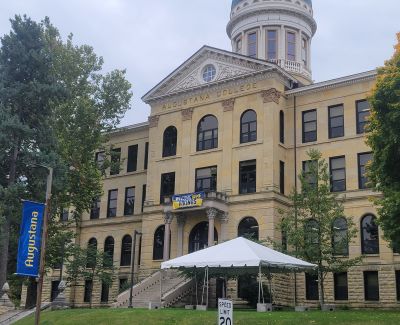 Front view of the main building at Augustana College, a large stone structure with 'AUGUSTANA COLLEGE' inscribed above the entrance, stairs leading up, and a white event tent on the lawn.
