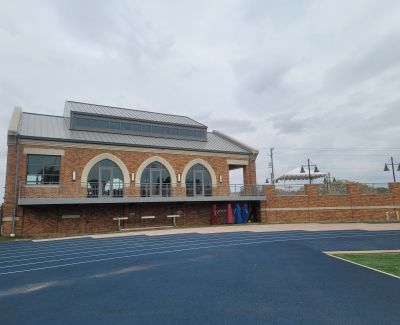 A modern brick building with large arched windows and a metal roof, possibly an athletic facility, overlooking a blue running track under a cloudy sky.