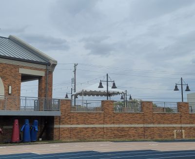  View of a brick wall and structure adjacent to a blue running track, with blue objects stored underneath an overhang and a white tent visible on an upper level in the background.