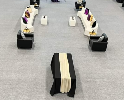 Overhead view of a modern event lounge area featuring curved white sofas and black armchairs arranged on grey carpet, accented with purple and gold pillows. A black-draped pedestal table is in the foreground.