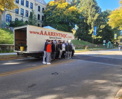 A group of people posing behind an open AAA Rents QC box truck parked on a road at Augustana College, with the domed main building and autumn trees in the background.