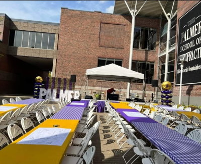 Outdoor event setup for Palmer School of Chiropractic, featuring long rows of tables with yellow and purple checkered cloths, white chairs, balloon decorations, and a small tent over a stage area.