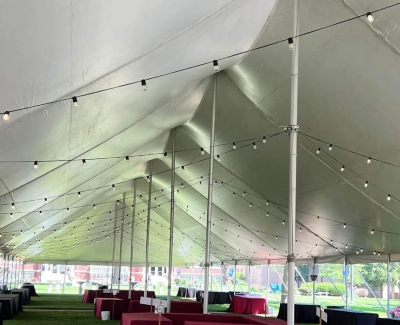 Interior view of a large white event tent showing strings of bistro lights hung beneath the canopy ceiling, illuminating tables with dark cloths set up on artificial turf below.