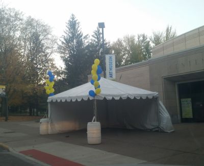 A white frame tent with grey sidewalls and blue/yellow balloon decorations set up outside the entrance to an Art Museum building.