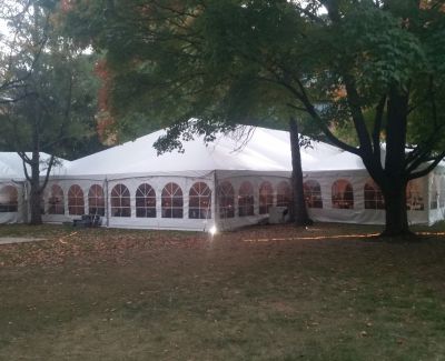 A large white frame tent with arched window sidewalls illuminated from within, pitched on a grassy area under trees during twilight or evening.