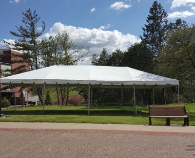 A long white frame tent erected on a grassy lawn next to a paved path and park bench, with trees and buildings in the background under a partly cloudy blue sky.
