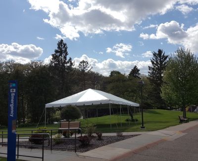 A white frame tent set up on a grassy knoll on a campus or park-like setting, near a paved walkway and a blue 
