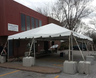 A white frame tent erected in a paved parking lot in front of a brick building, secured with large concrete block weights at each leg.