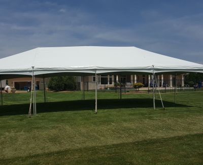 A long, rectangular white frame tent set up on a well-manicured green lawn under a partly cloudy sky, secured with stakes, with a building visible in the distance.
