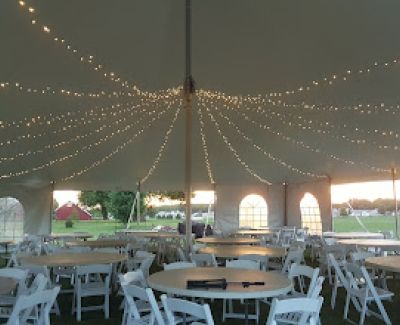 Interior view from under a large event tent looking up at strings of bistro lights crisscrossing beneath the white canopy, with round tables and white chairs set up for guests below.